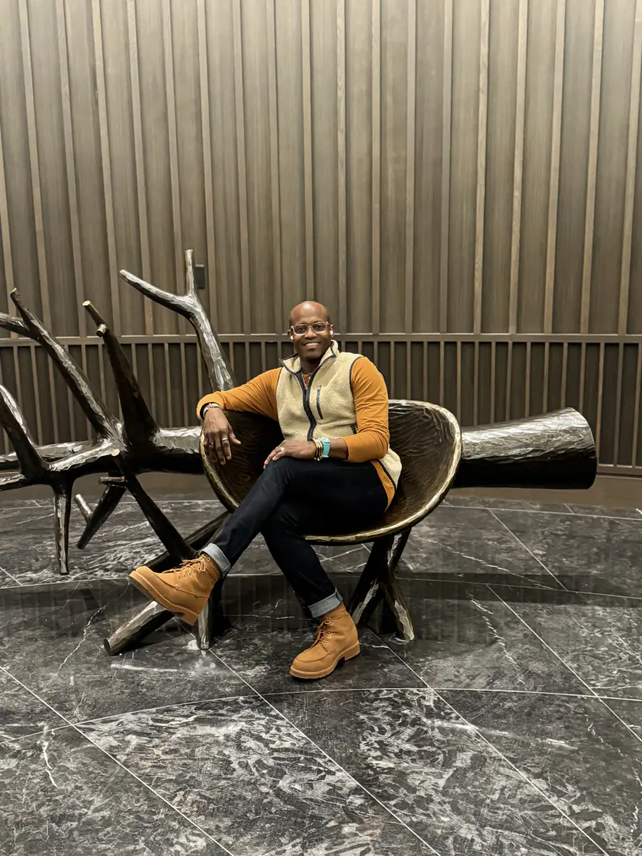 Maurice Howell sits beside a sculptural chair in a wood-paneled interior.