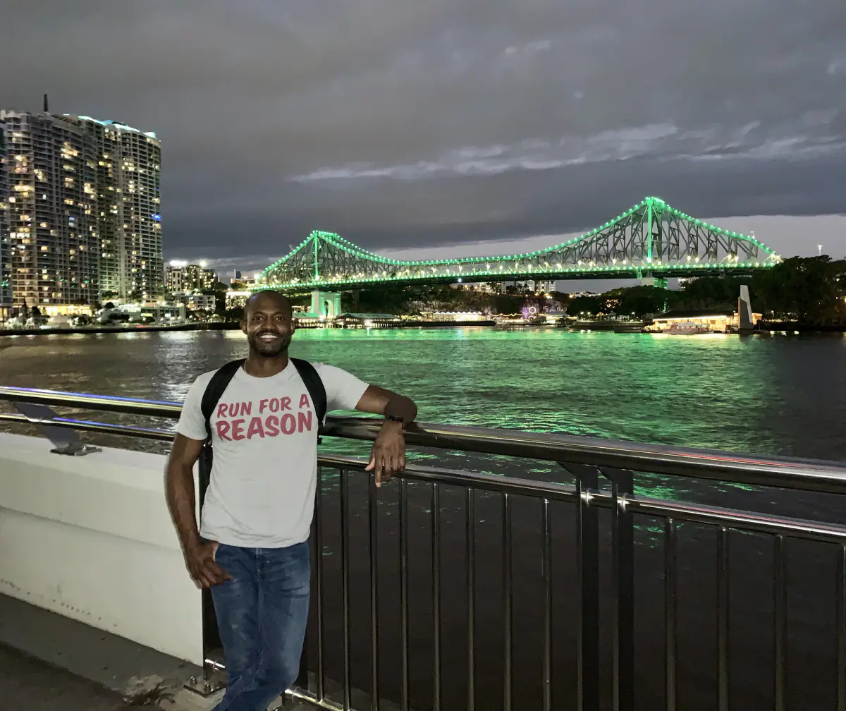 Maurice Howell stands by a waterfront railing with a green-lit steel bridge behind him at night.