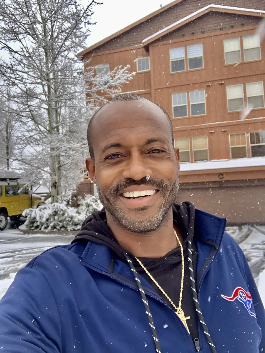 Maurice Howell smiles outdoors as snow falls, with trees and a building behind him.