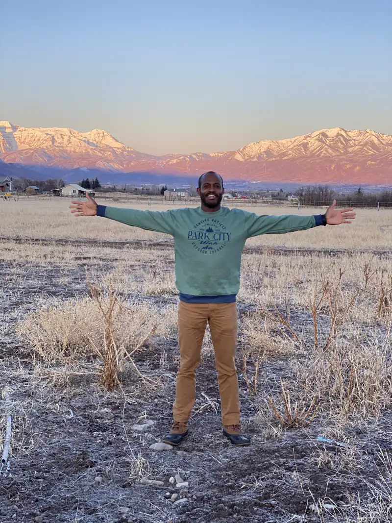 Maurice Howell stands with his arms open in a field with snow-covered mountains behind him.