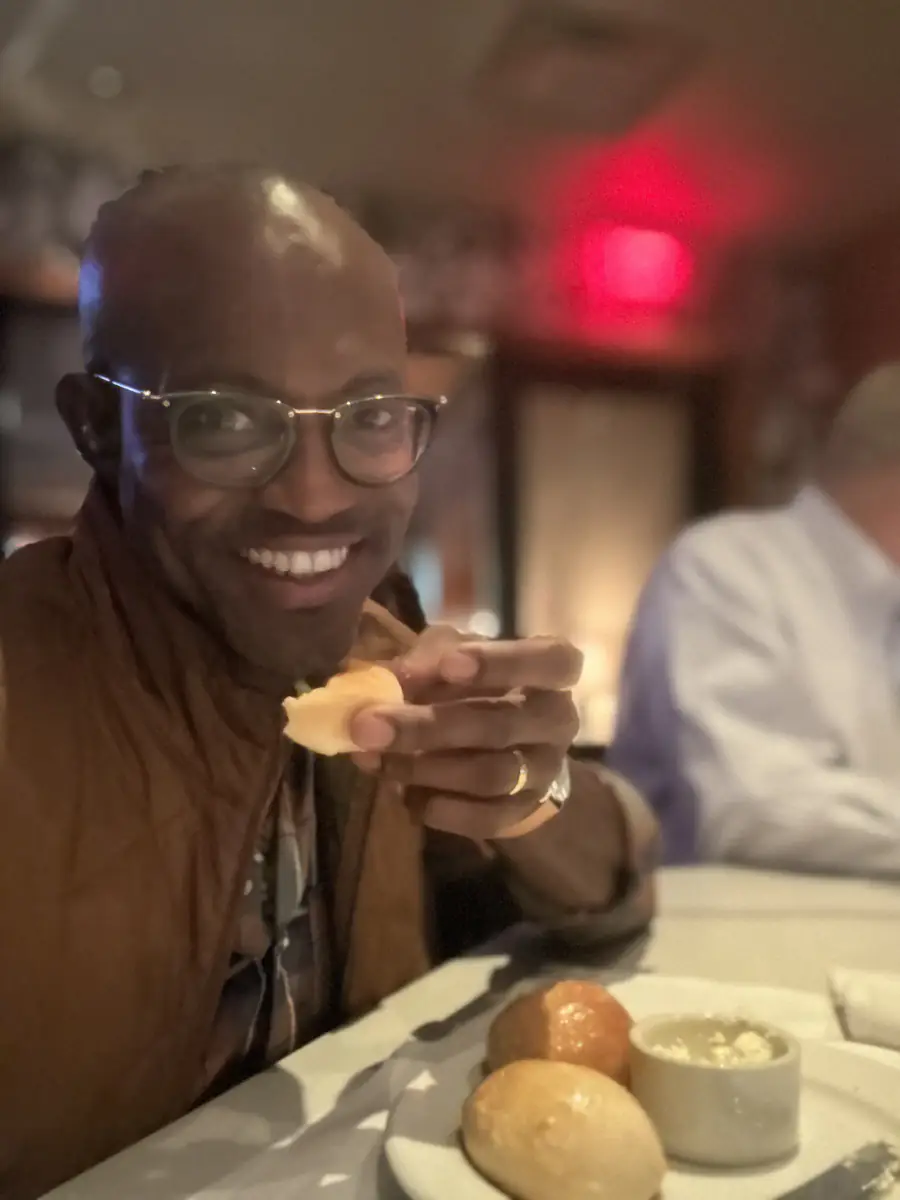Maurice Howell smiles and holds bread at a restaurant table, with another diner out of focus.